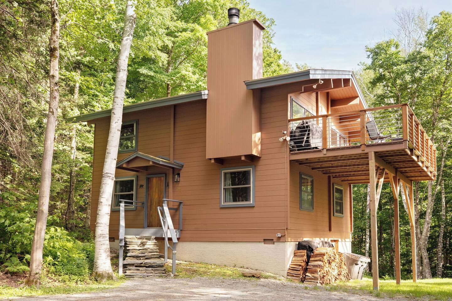 A two-story wooden cabin with a balcony and a chimney, surrounded by trees.