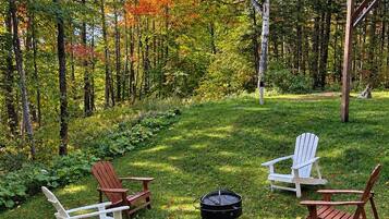 A forested area with a deck and three Adirondack chairs arranged around a fire pit.