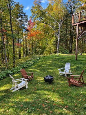 Een bebost gebied met een terras en drie Adirondack-stoelen rond een vuurkuil.