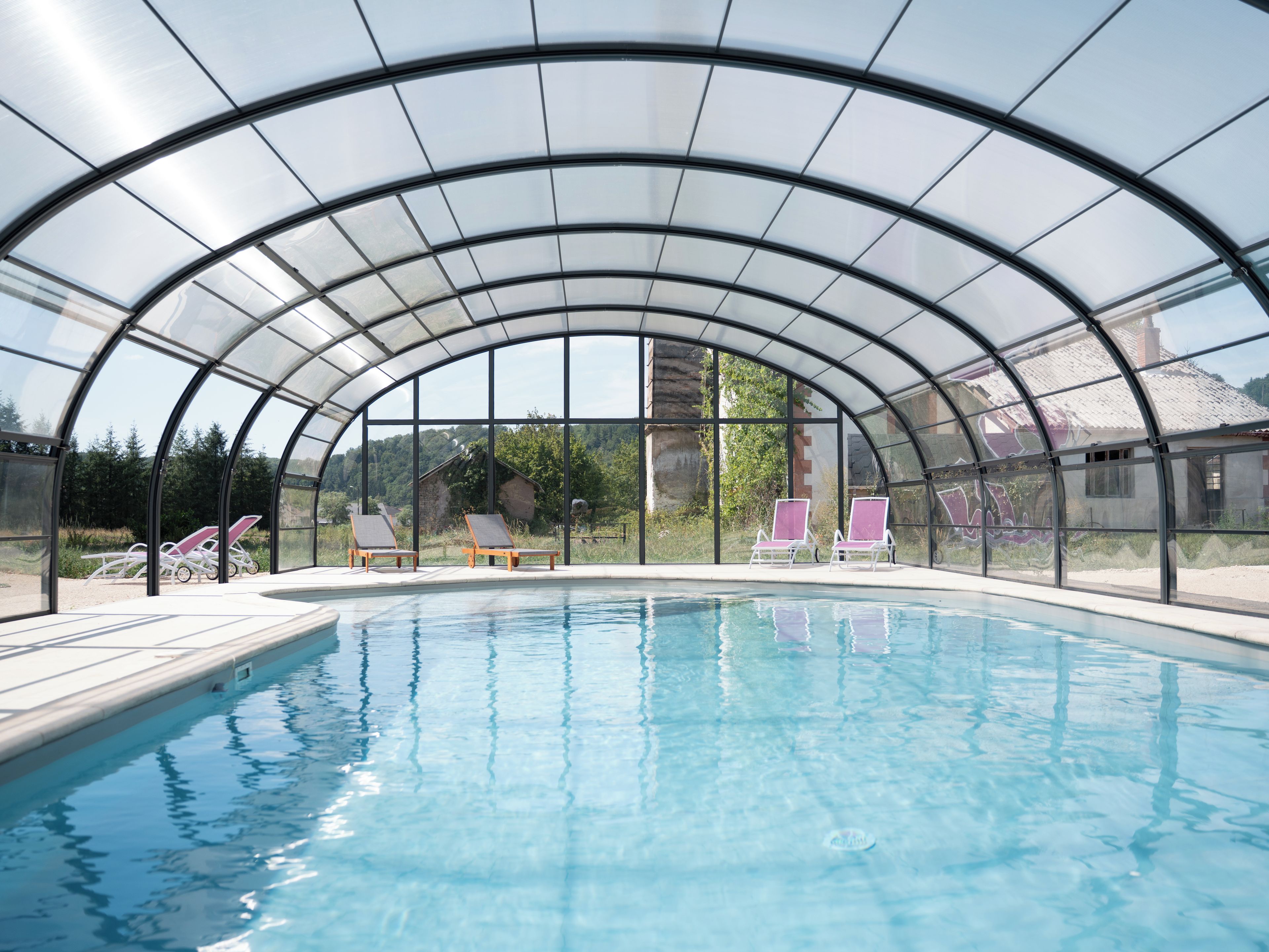 Une piscine couverte avec des parois en verre transparent, entourée de chaises longues.