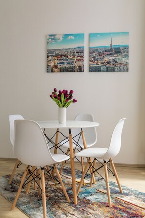 A dining area with a white table, four white chairs, and a vase with purple flowers. There are two cityscape paintings on the wall. - 1-Bedroom Apartment | Naschmarkt | 4 Guests | Self Check-in (Wien)