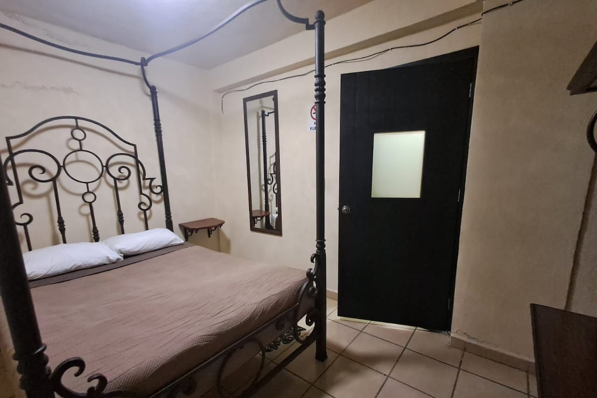 A bedroom with a four-poster bed, a small bedside table, a mirror, and a door with a window.