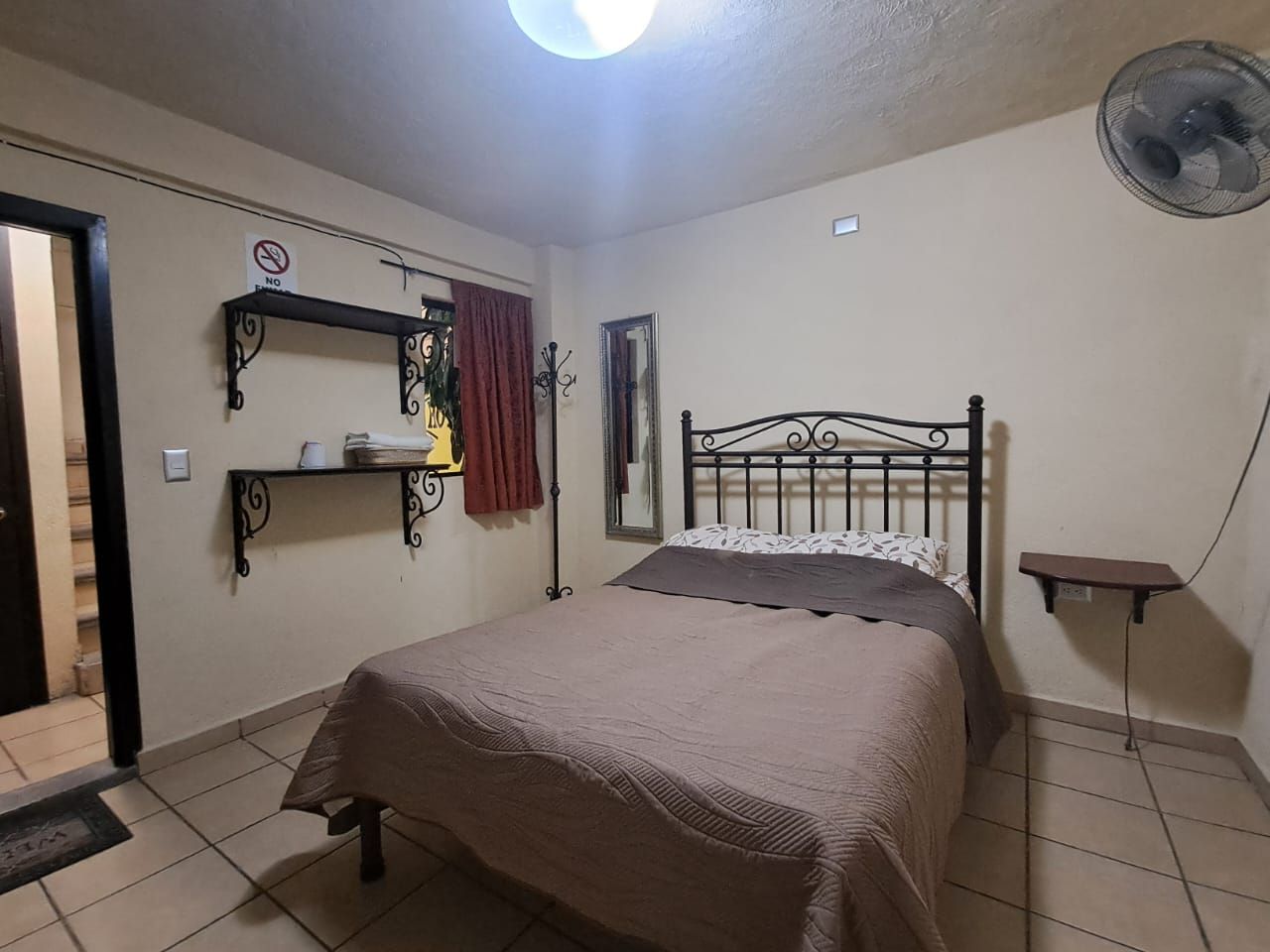 A bedroom with a bed, a shelf with a lamp, a mirror, and a fan.