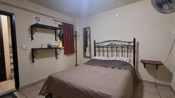 A bedroom with a bed, a shelf with a lamp, a mirror, and a fan.