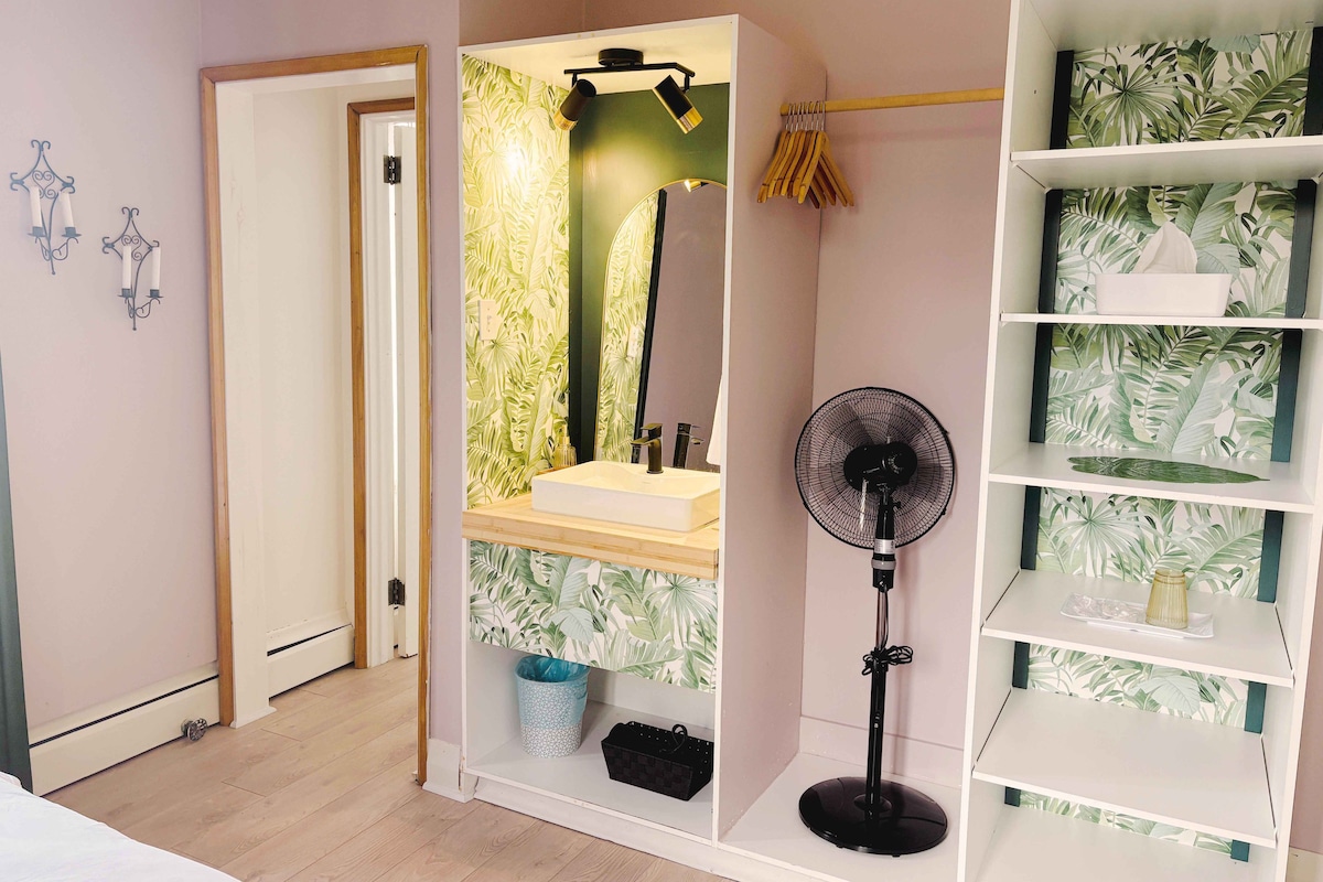A bedroom with a bed, a bedside table, a mirror, a fan, a shelf with decorative items, and a door leading to another room.