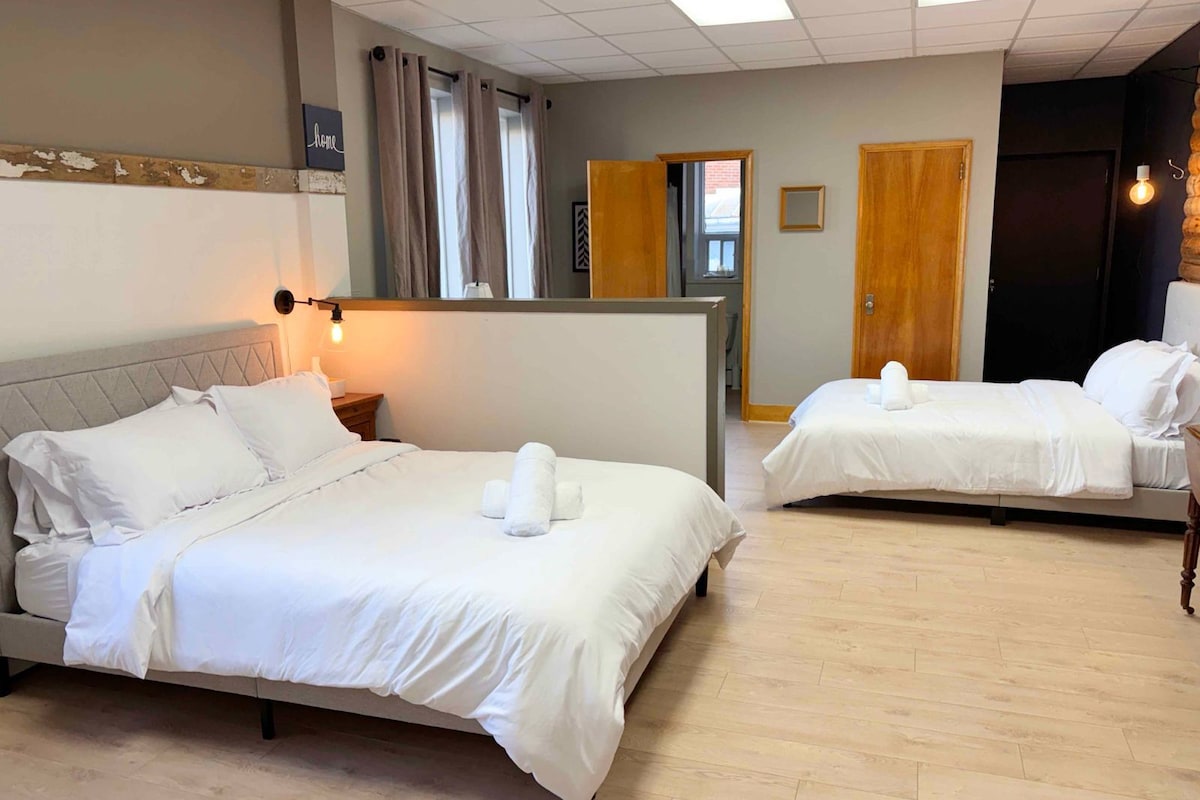 Une chambre d’hôtel avec deux lits, une table de chevet, une lampe, une fenêtre avec des rideaux et une porte.