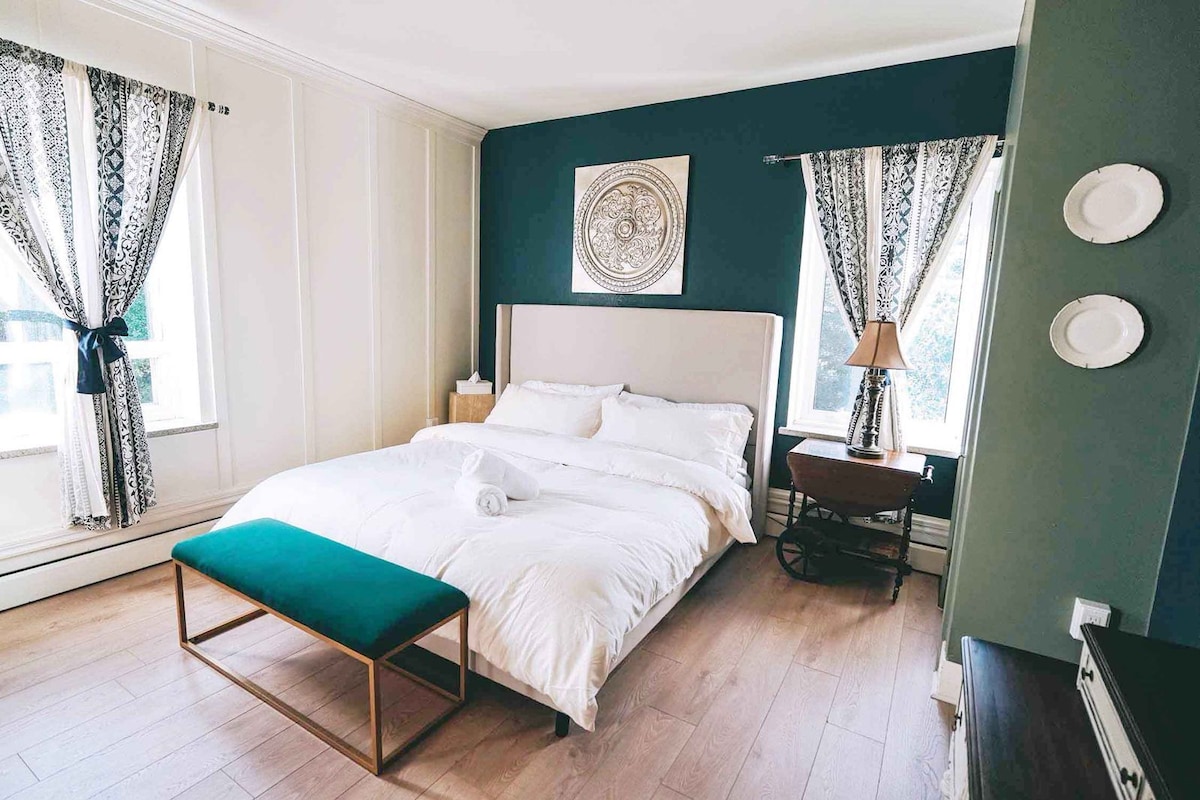 Un lit bien fait, avec du linge de lit blanc, un banc de couleur turquoise et une table de chevet avec une lampe.