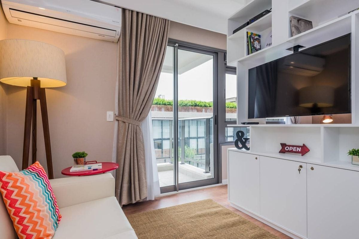 Ein modernes Wohnzimmer mit einer Couch, einem Fernseher, einem kleinen Tisch mit einer Lampe und einer Glastür, die auf einen Balkon führt.