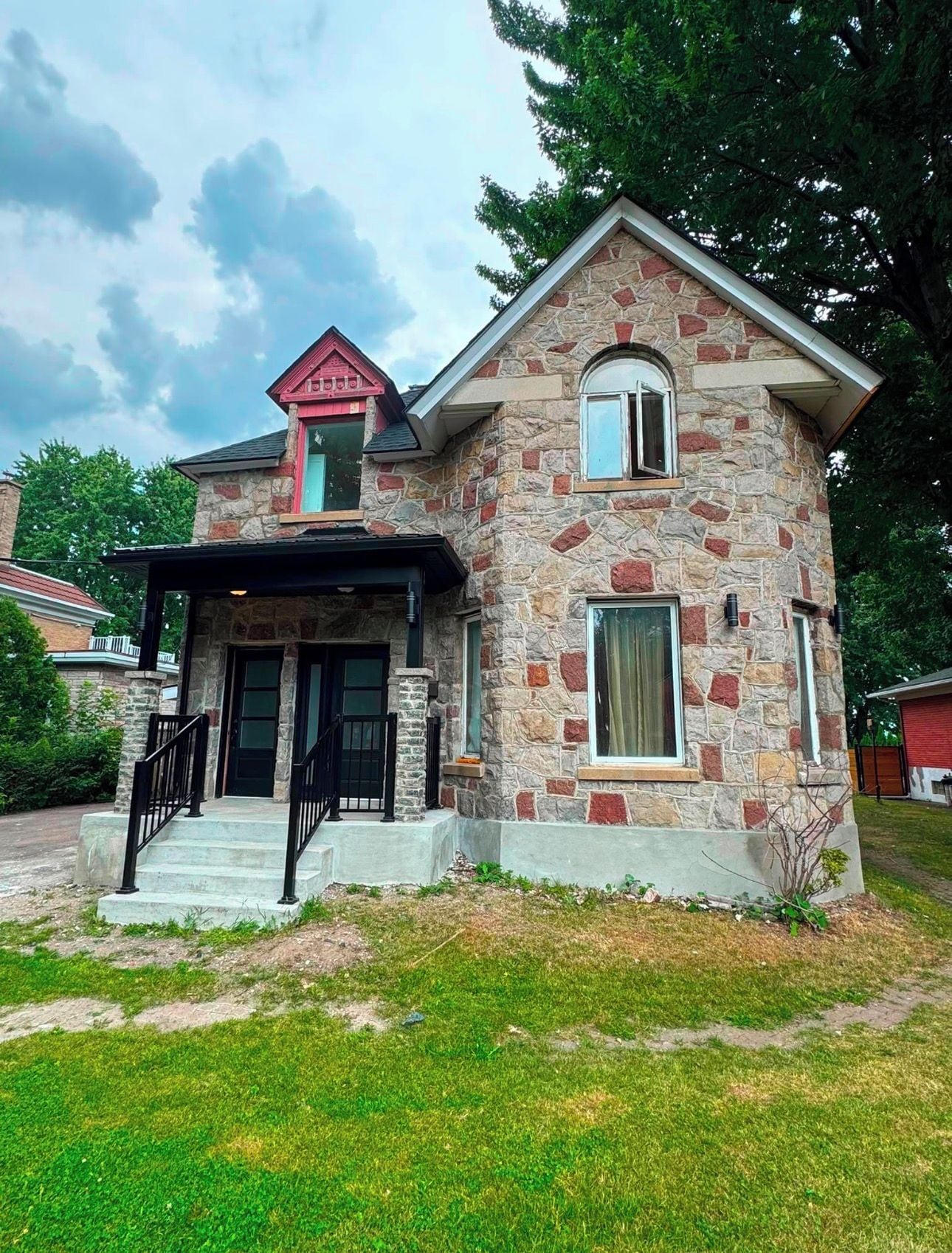A two-story stone house with a red roof, a front porch, and a small garden area.