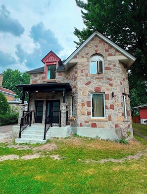 Ein zweigeschossiges Steinhaus mit rotem Dach, einer Veranda und einem kleinen Garten.