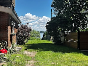 Ein Wohnhof mit einem Ziegelhaus, einem Holzzaun und einem grasbewachsen Weg, der zu einem Gewässer führt.