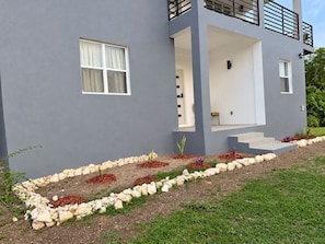 A modern two-story house with a grey exterior, a visible staircase leading to the upper floor, and a landscaped garden with a variety of plants and a stone border. - Zu Casa | Peaceful 2-Bedroom Apartment in Historic Parham, Antigua (Saint Peter’s)