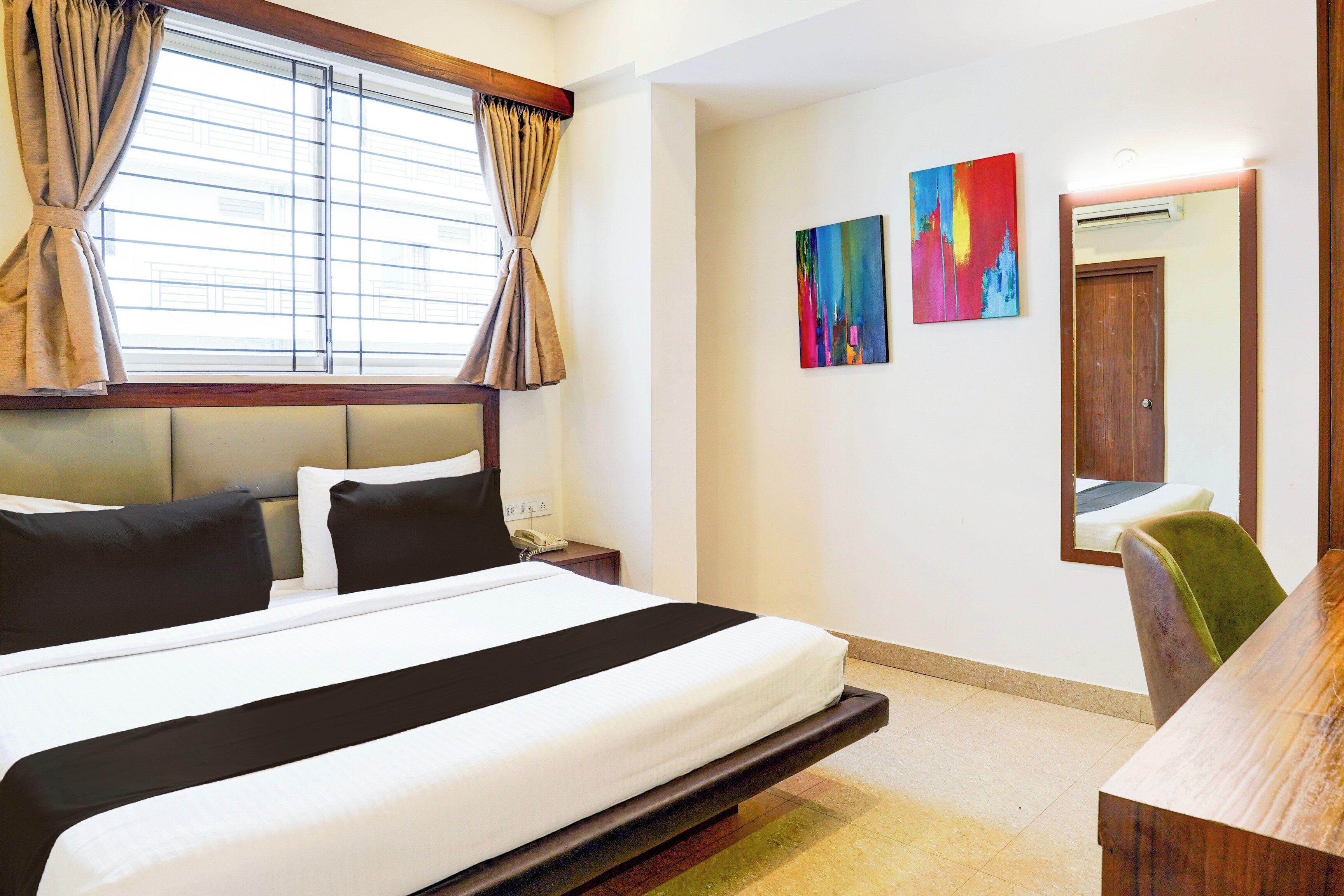 Habitación de hotel con cama, mesita de noche, teléfono, espejo y dos cuadros abstractos en la pared.