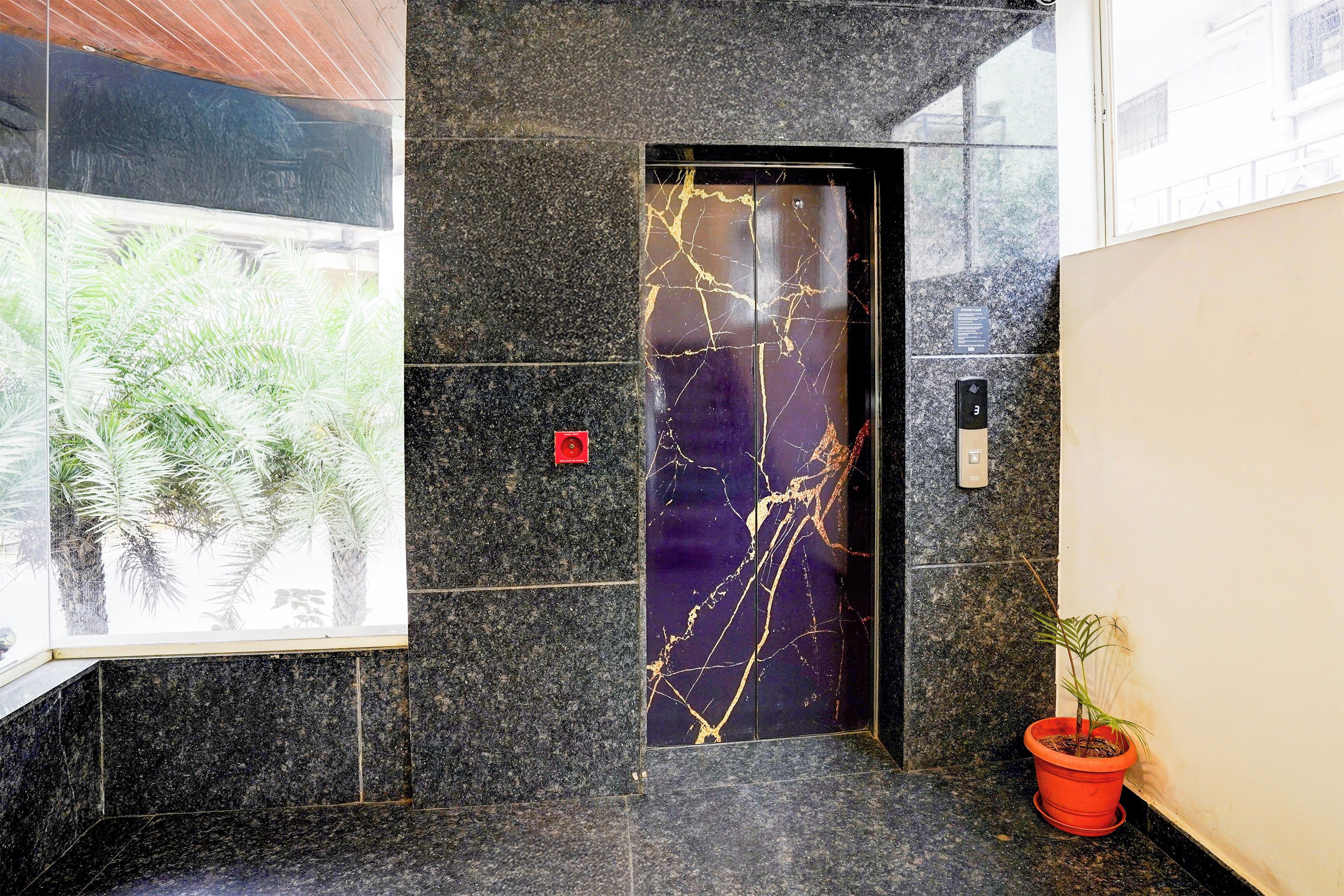 A marble elevator door with a reflective surface, a red emergency button, and a potted plant beside it.