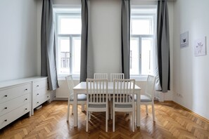 A room with a dining table and chairs, a white dresser, and large windows with curtains. - 2-Bedroom Apartment with Elevator | Naschmarkt | Sleeps 6 (Wien)