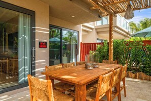 An outdoor patio with a wooden table and chairs, a glass door with a 'NO SMOKING' sign, and a view of greenery. - 2BR Luxury Escape | Pool, Palapa & Outdoor Shower (Noord)