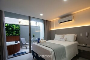 Ein Hotelzimmer mit einem Bett, einem Schreibtisch, einem Sessel, einem kleinen Tisch, einer Pflanze und einer Klimaanlage.