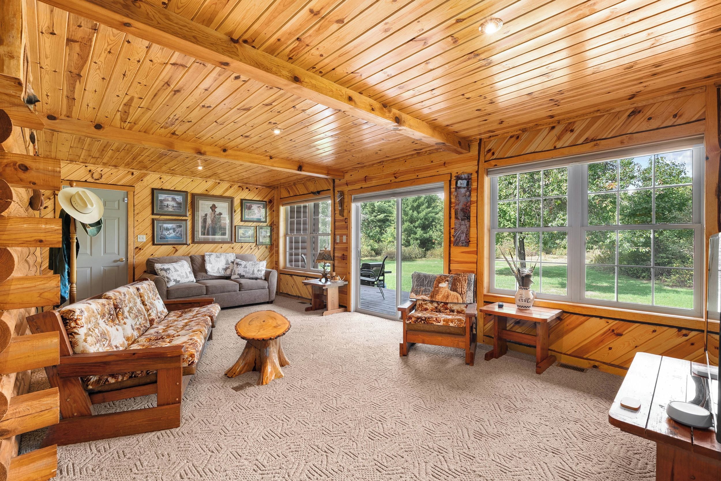 A spacious room with wooden walls and ceiling, a sofa set, a coffee table, and a view of a green landscape through large windows.