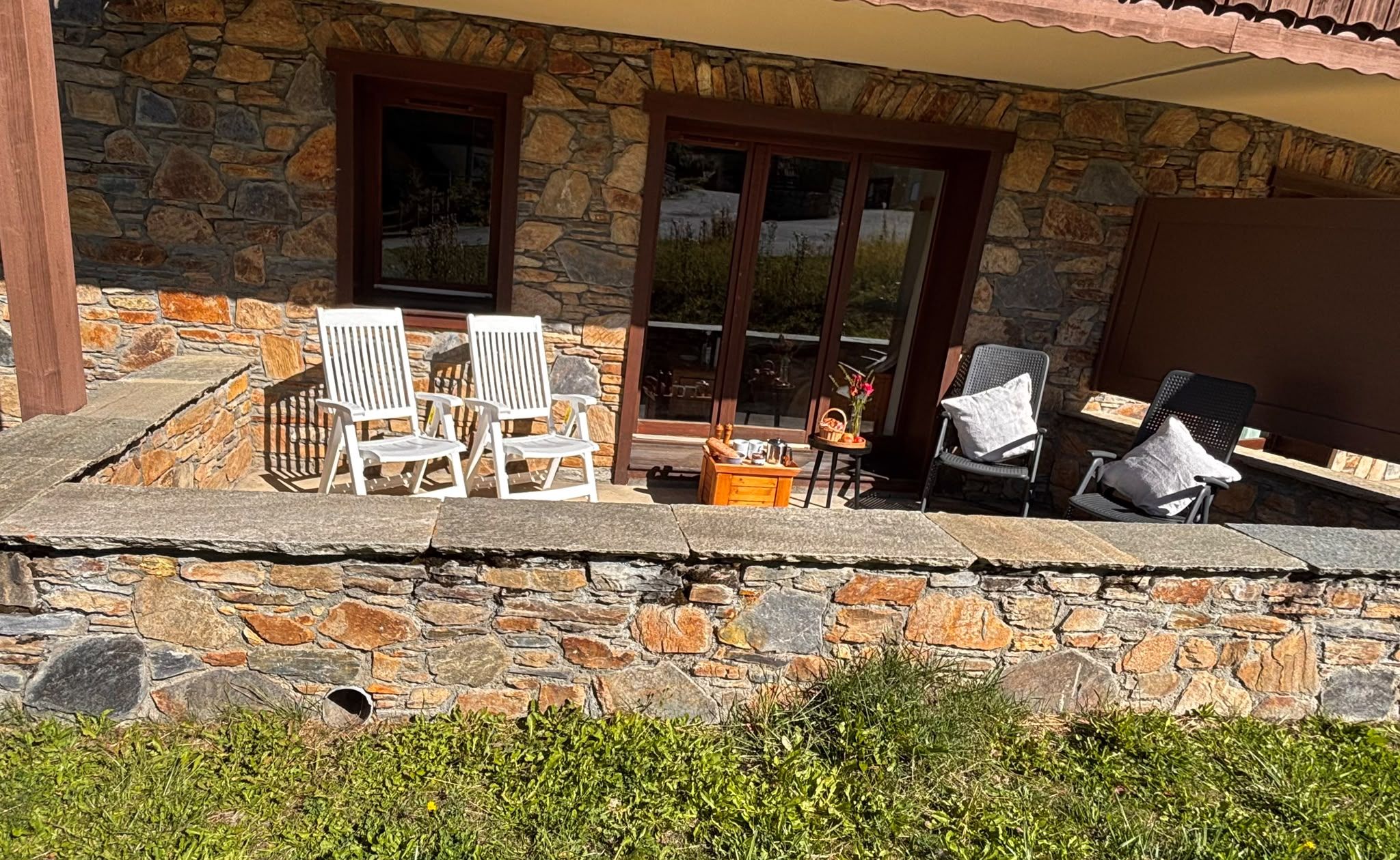 A stone house with a patio area featuring white chairs, a table, and a small wooden table with items on it.