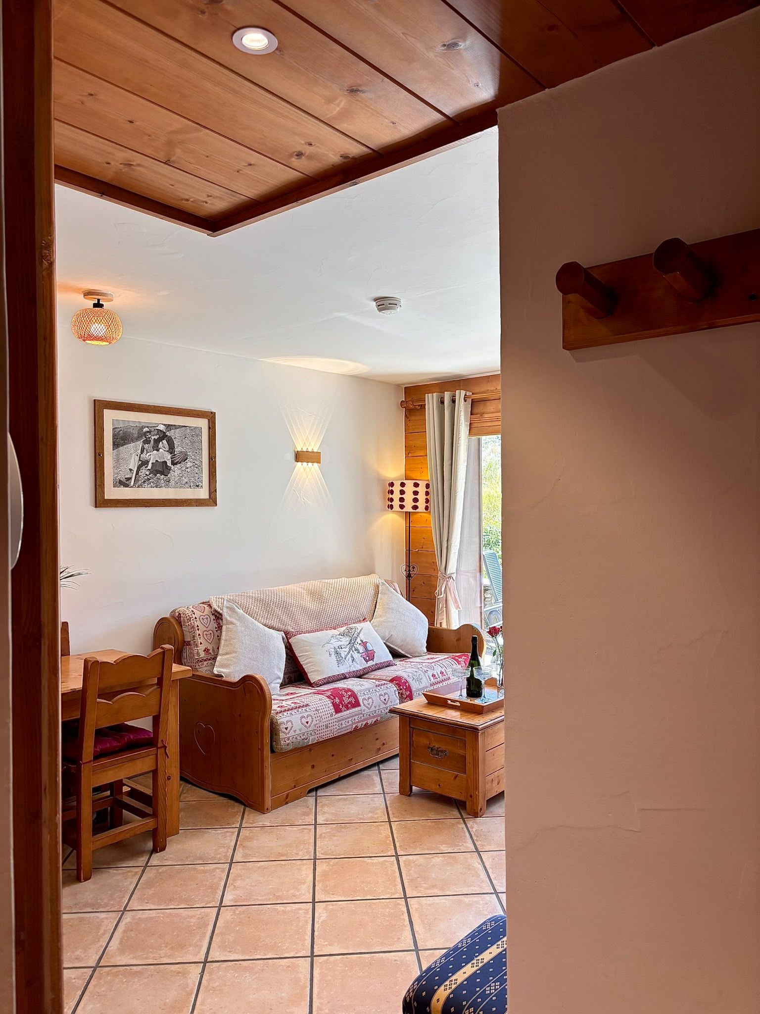 A cozy room with a wooden ceiling, a bed with patterned bedding, a wooden table with a bottle, and a wall-mounted light fixture.