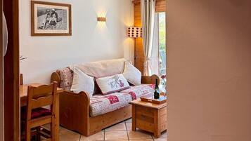 A cozy room with a wooden ceiling, a bed with patterned bedding, a wooden table with a bottle, and a wall-mounted light fixture.
