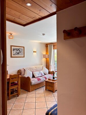 A cozy room with a wooden ceiling, a bed with patterned bedding, a wooden table with a bottle, and a wall-mounted light fixture.