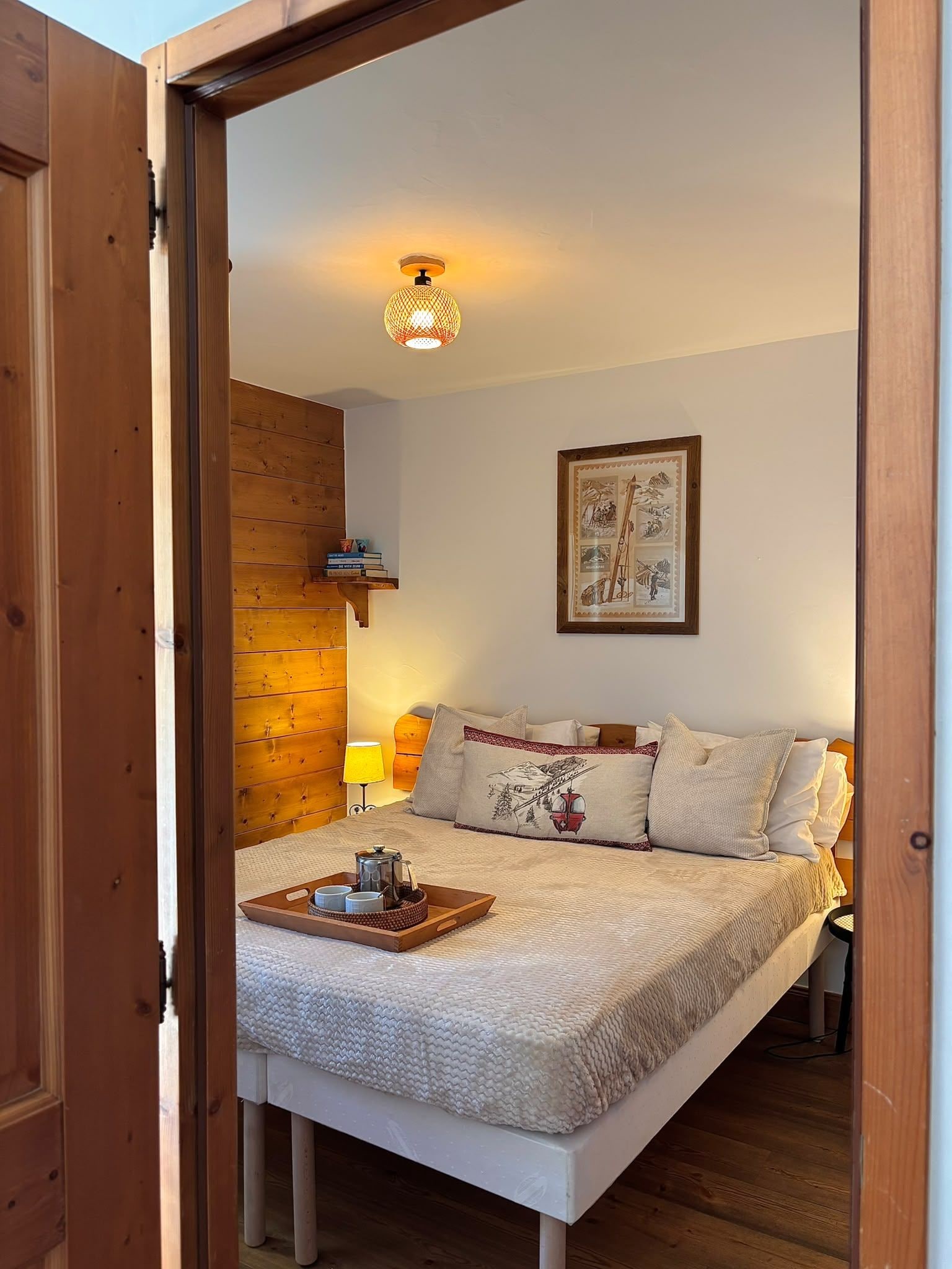 A bedroom with a bed, pillows, a bedside table with a tray, a lamp, and a wall-mounted shelf with books.