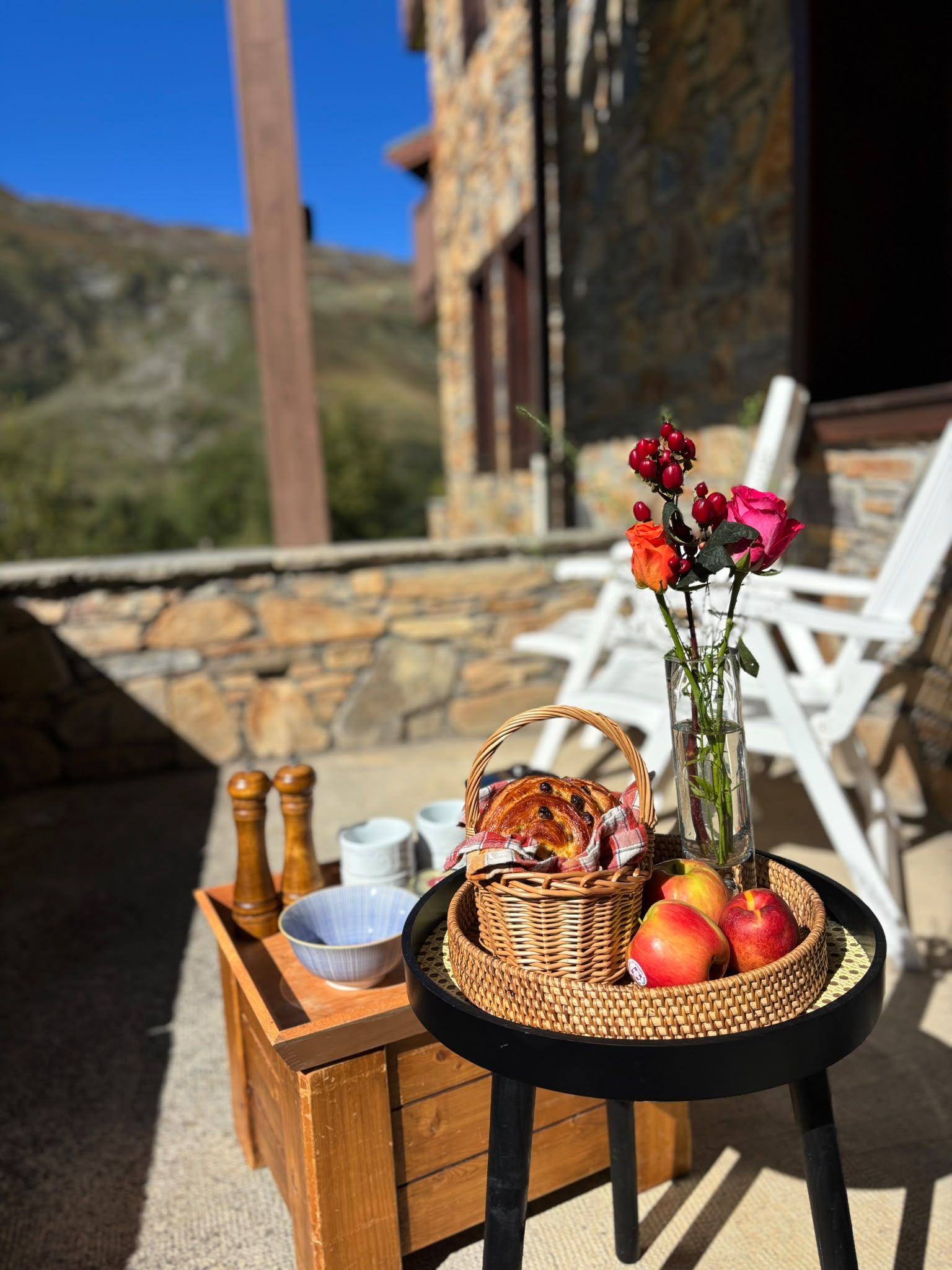 Et bord med en kurv med brød, æbler og en vase med blomster, placeret på en terrasse med stenvægge og hvide stole.