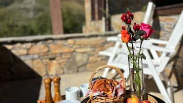 Et bord med en kurv med brød, æbler og en vase med blomster, placeret på en terrasse med stenvægge og hvide stole.