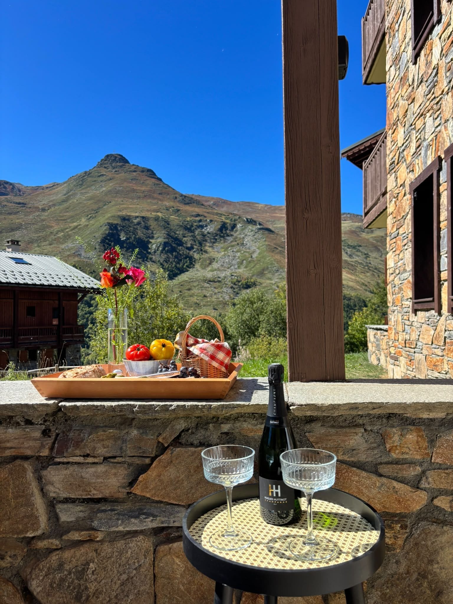 A table with a bottle of champagne and glasses, a basket with food, and a flower arrangement, set against a mountainous backdrop.