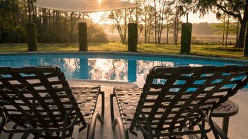 A poolside area with two lounge chairs and a table, surrounded by trees and a clear sky.