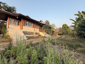 Una casa de madera rústica con base de piedra, un porche y un jardín con diversas plantas y un árbol de plátano.