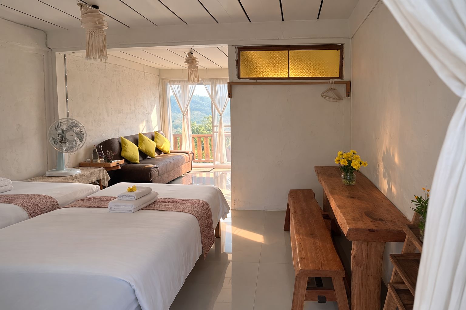 A bedroom with two beds, a wooden bench, a table with flowers, and a fan.