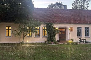 A two-story building with a tiled roof, multiple windows, and a front door. There is a small garden area with grass and a few plants. - Cozy 2-room apartment for a relaxing getaway (Müden (Aller))