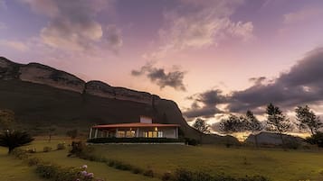 Uma casa moderna com telhado plano, rodeada por um gramado bem cuidado e uma grande montanha ao fundo.