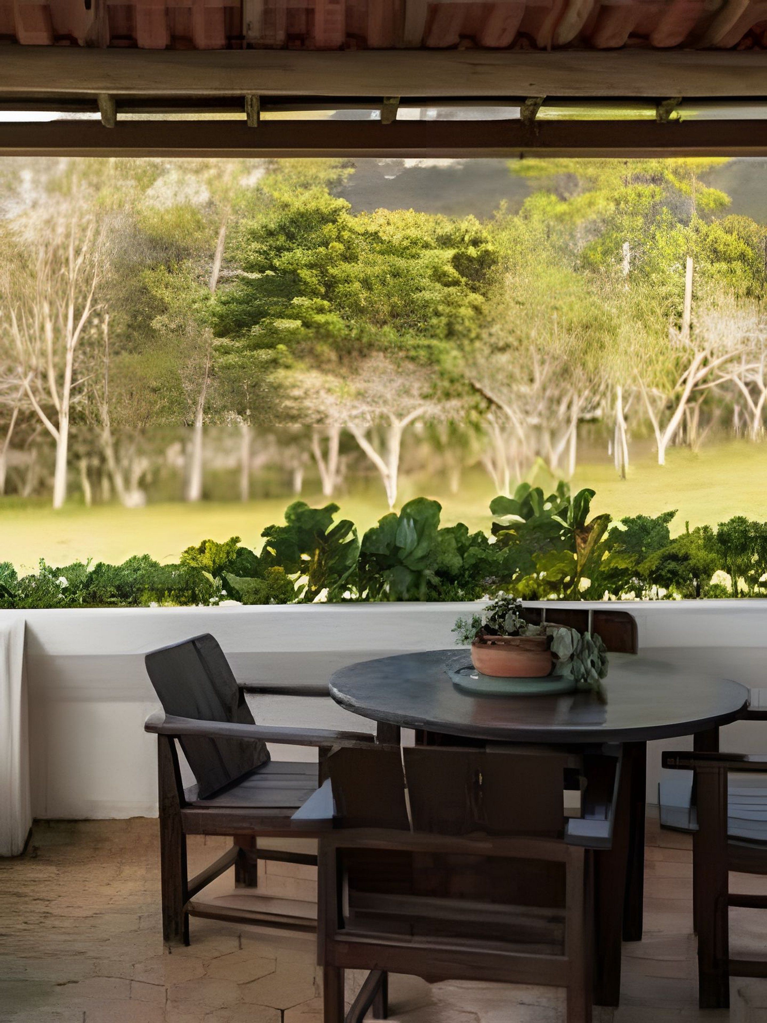 Uma área de estar coberta ao ar livre, com mesa e cadeiras, oferece vista para uma paisagem verdejante com árvores e um campo gramado.