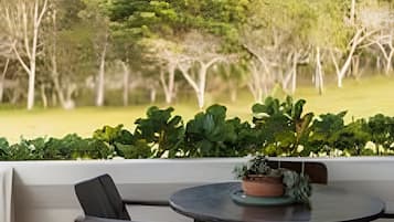 Uma área de estar coberta ao ar livre, com mesa e cadeiras, oferece vista para uma paisagem verdejante com árvores e um campo gramado.