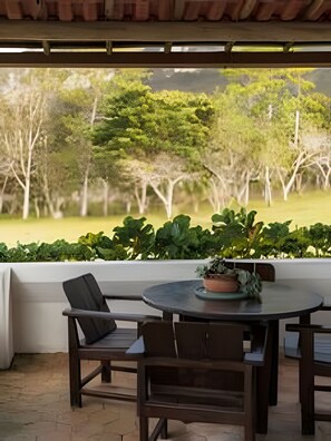 Un área de estar al aire libre techada con una mesa y sillas, con vistas a un paisaje verde exuberante con árboles y un campo cubierto de césped.