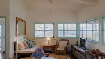 Quarto com mesa de jantar de madeira, sofá com almofadas coloridas, cadeira, mesinha com luminária, televisão e janelas amplas com vista para o mar.