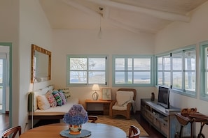 A room with a wooden dining table, a sofa with colorful cushions, a chair, a small table with a lamp, a television, and large windows with a view of the sea.