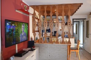Una sala de estar con televisión, un separador de madera y una zona de cocina al fondo.