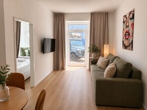 A modern hotel room with a sofa, a TV, a dining area with a table and chairs, and a view of the street through a glass door. - 1-Bedroom Apartment with Air Conditioning | Danube Canal | 4 Guests (Wien)