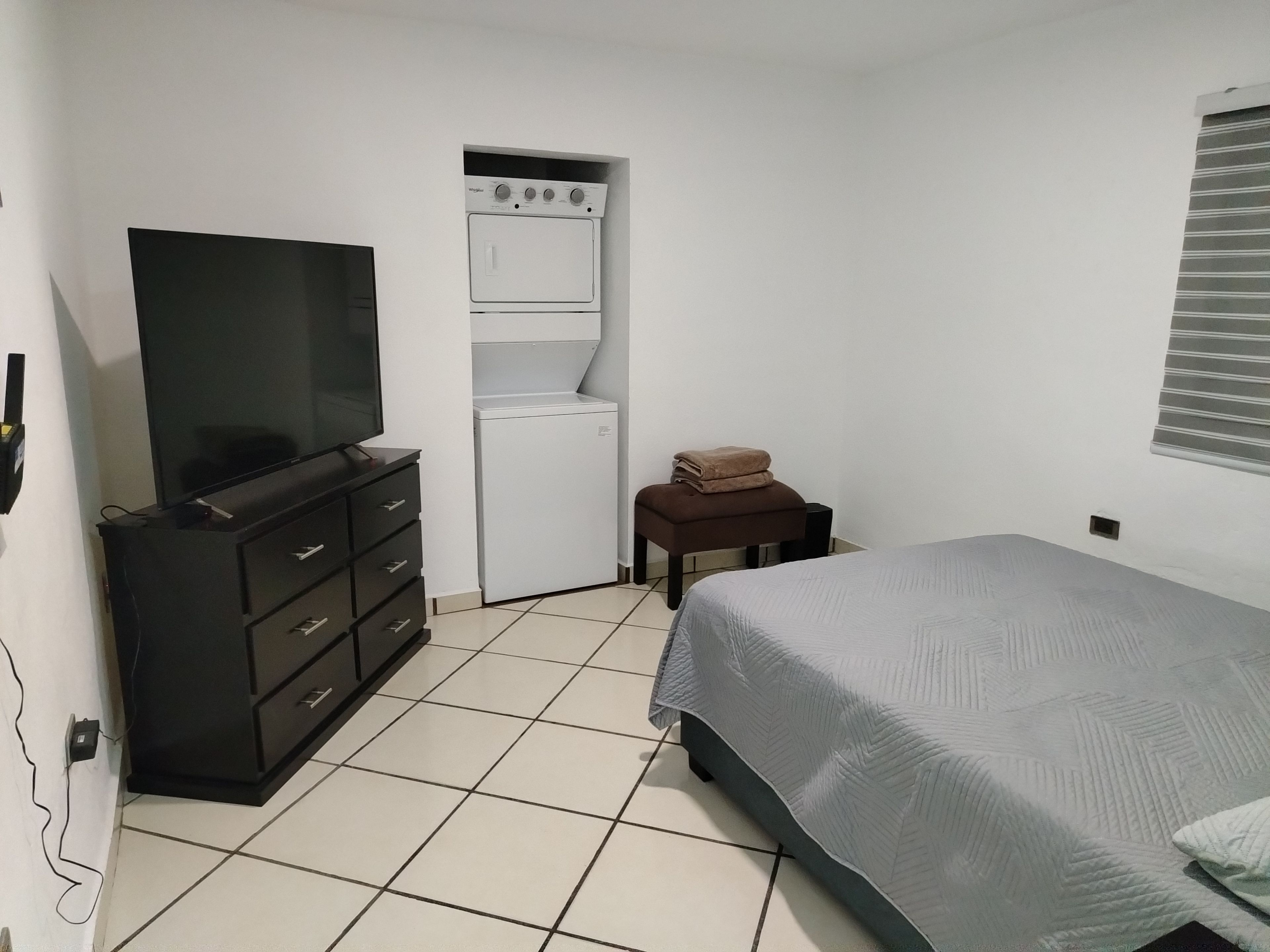 A bedroom with a bed, a television on a dresser, a washing machine, and a small stool.