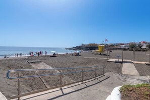 Ein Strand mit einem Metallgeländer im Vordergrund, einem Sandstrand und Gebäuden im Hintergrund.