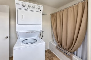 A laundry room with a front-loading washer and dryer, a window with beige curtains, and a tiled floor. - Rest Easy in Dildo | By the Sea | South Dildo (South Dildo)