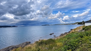 Paisaje costero con costa rocosa, mar tranquilo y una masa de tierra distante bajo un cielo parcialmente nublado.