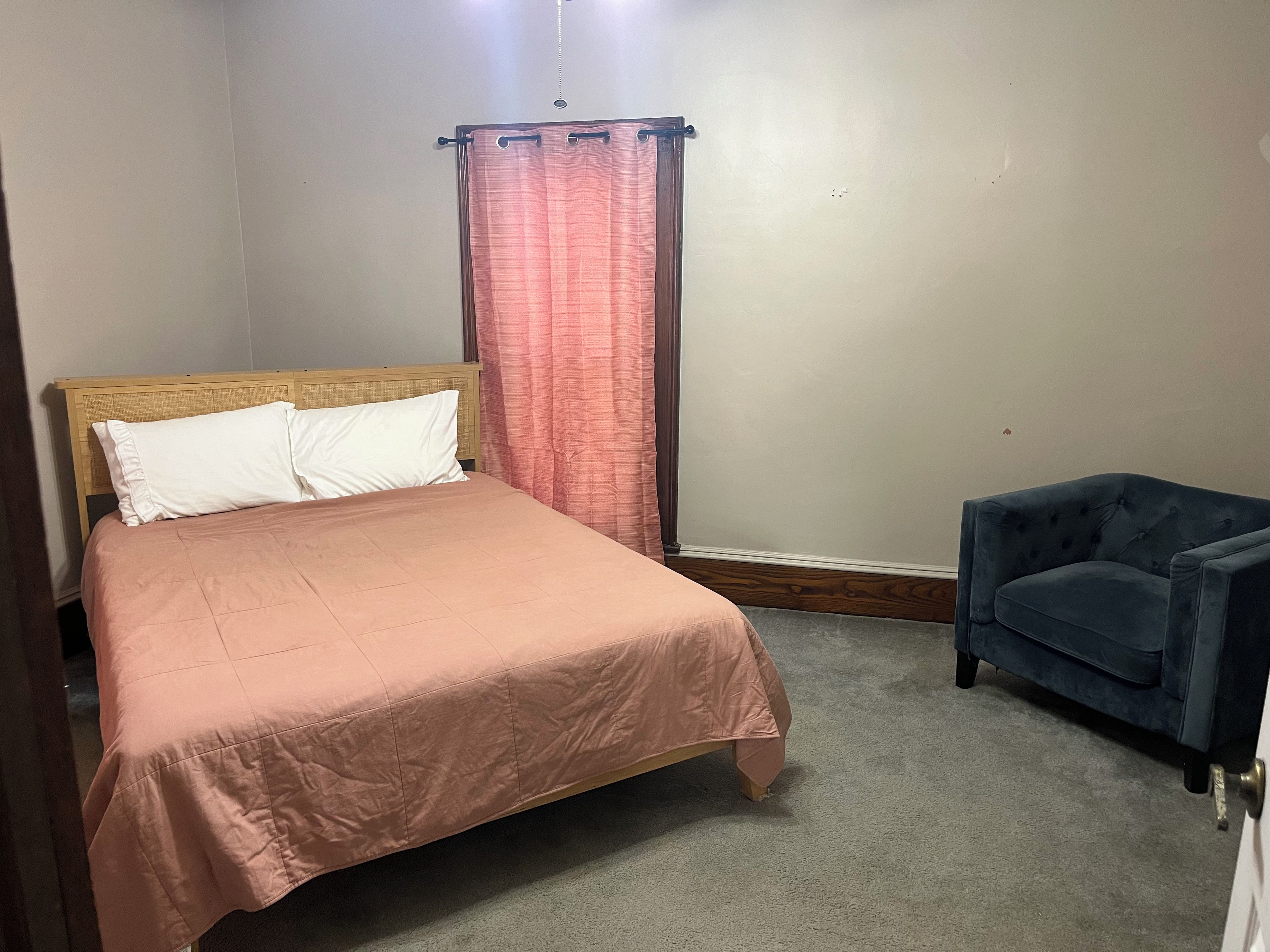 Un lit simple avec une tête de lit en bois, un fauteuil bleu et un rideau rose.