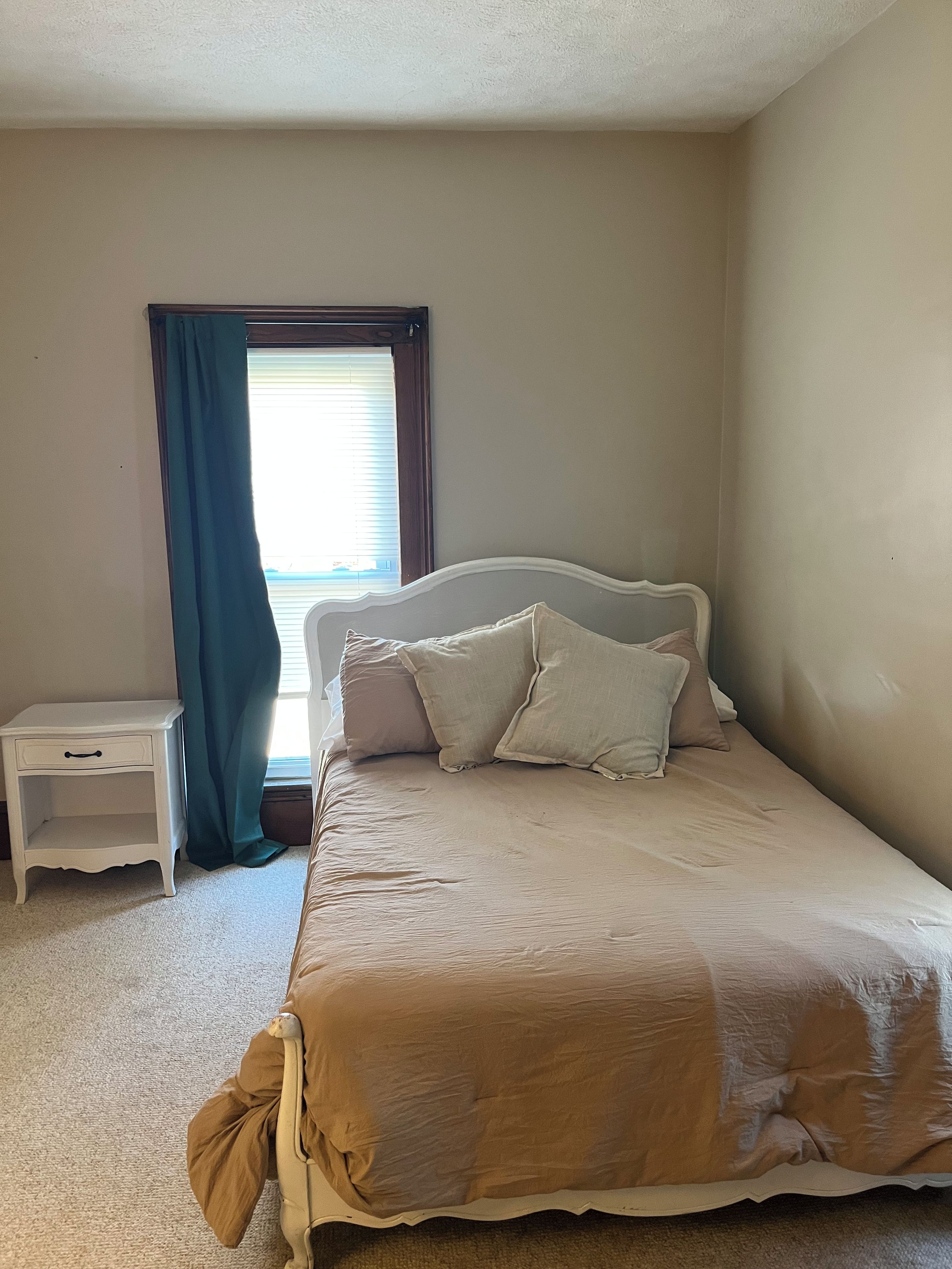 Une chambre à coucher avec un lit, une table de chevet, une fenêtre avec des rideaux et un sol recouvert d’une moquette.