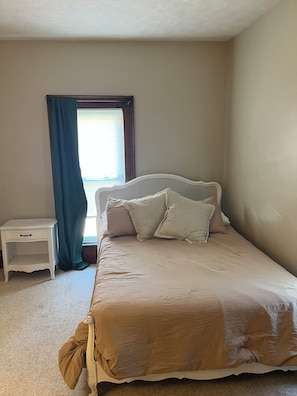 Une chambre à coucher avec un lit, une table de chevet, une fenêtre avec des rideaux et un sol recouvert d’une moquette.