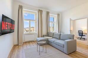 A modern living room with a grey sofa, a small side table, a flat-screen TV mounted on the wall, and a window with curtains. - 1-Bedroom Apartment with Air Conditioning | Landstraße | 4 Guests (Wien)
